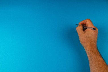 Bir el, parlak mavi bir arka plana karşı bir kalemi kavrıyor, düşünceleri ya da fikirleri not etmeye hazır. Sahne yaratıcı ve odaklanmış, yazmak için mükemmel..