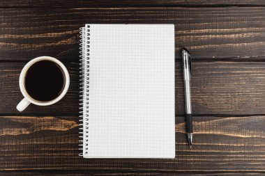 Şebeke desenli boş bir defter, siyah bir kahve fincanı ve ahşap bir masada gümüş bir kalemle yan yana durur. Not almak ya da planlamak için mükemmel bir ayar.