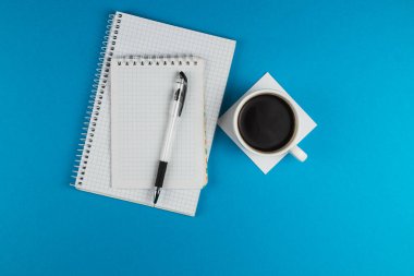 Katı mavi bir yüzeye yerleştirilmiş iki defter, bir kalem ve bir kahve fincanı içeren bir çalışma alanı düzeneği. Çalışmak ya da planlamak için mükemmel.