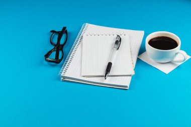 Bir spiral defter, kalem ve bardaklar parlak mavi bir masanın üstünde, dumanı üstünde bir fincan kahvenin yanında otururlar. Bu sahne bir rahatlama anı yakalar ve odaklanır..