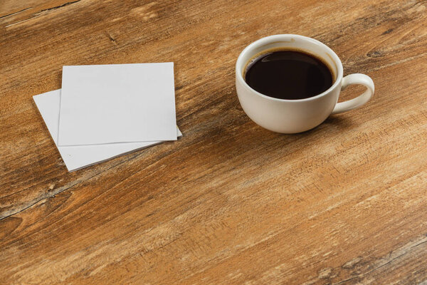 A white coffee cup filled with dark coffee sits on a rustic wooden table. Next to it are two blank notes, creating a relaxed and inviting atmosphere.