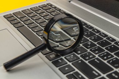 Bir büyüteç dizüstü bilgisayarın klavyesine yerleştirilir. Sahne parlak renkler içeriyor ve anahtarların detaylarına odaklanıyor, çalışma alanı atmosferini artırıyor.