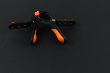 Turuncu saplı siyah bir klemp aleti karanlık bir yüzeyde duruyor. Tasarımı kolay kullanım ve güvenli klempleme olanağı sağlıyor. Çeşitli atölyeler ve projeler için ideal.