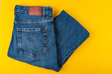 Bir çift mavi kot pantolon düzgünce katlanmış ve parlak sarı bir yüzey üzerine serilmiş, klasik stil ve renk kontrastını minimal bir ayarda sergiliyor.