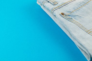 Katlanmış açık mavi kotlar canlı bir turkuaz arka planda güzelce düzenlenir, kot kumaşı ve renk kontrastını basit ama çekici bir şekilde vurgular..