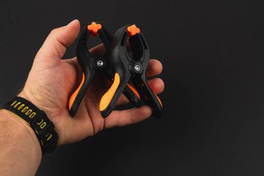 Bir el, iki siyah ve turuncu kıskacı koyu arka planda tutuyor. Kıskaçlar nesneleri güvenli bir şekilde bağlamak ya da tutmak için tasarlanmıştır..