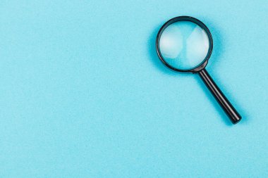 Bir büyüteç pürüzsüz mavi bir yüzeye dayanır, altındaki nesnelerin detaylarını merak uyandırır ve araştırır..