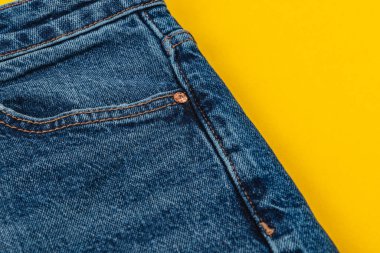 Mavi kot kumaşın yakın plan görüntüsü dikişler ve cep gibi ayrıntıları parlak sarı arka planda, moda ve tekstil tasarımı vurgulanıyor.