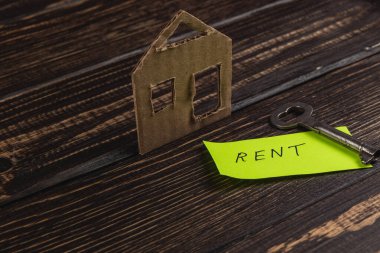 Kartondan bir ev modeli, kiralık bir anahtarın yanında duruyor. Koyu ahşap bir masaya yerleştirilmiş parlak bir not. Kiralık bir emlak temasını işaret ediyor..
