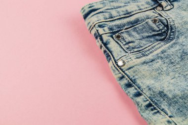 Denim jeans lie on a soft pink surface, highlighting the back pocket. The fabric is lightly worn, with a classic blue shade and shiny metal studs.