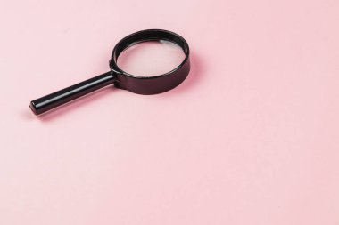 A black magnifying glass rests on a light pink surface, ready for close examination of details. The scene is simple and visually appealing, inviting curiosity.