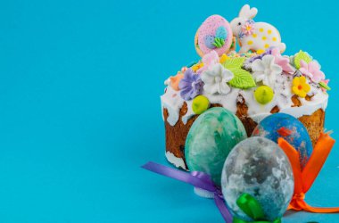 Kremalı bir pasta mavi bir yüzeye oturur. Pastanın yanında renkli yumurtalar var. Sahnede bahar ve Paskalya kutlaması için dekorasyonlar gösteriliyor..