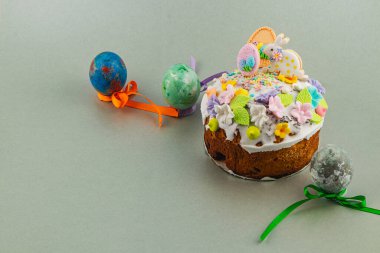 Yuvarlak bir pasta renkli krema ve çiçeklerle süslenmiş gri bir yüzeye oturur. Yakınlarda, üç tane süslü yumurta var. Kurdelelerle bağlanmış, Paskalya temalarını gösteriyor..