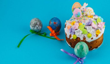 Üzerinde krema olan bir pasta mavi bir yüzeye yerleştirilir. Pastanın yanında kurdeleli renkli yumurtalar var. Ayar, Paskalya için neşeli bir ruh hali gösteriyor..