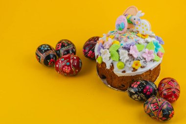 Bir pasta çeşitli malzemelerle süslenir ve parlak sarı bir masaya oturur. Yakınlarda, renkli tasarımlar ve desenler gösteren çeşitli boyalı yumurtalar sergileniyor..