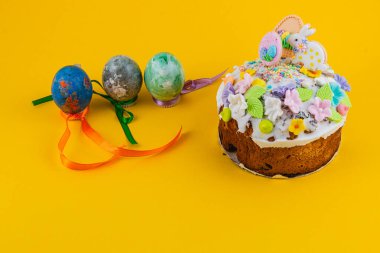 Renkli süslemeleri olan yuvarlak bir pasta masanın üzerinde oturur. Yanında üç tane kurdeleli Paskalya yumurtası var. Sahne, parlak sarı bir arkaplanda geçiyor..
