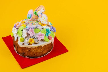 Renkli krema, çiçek ve tavşan figürleriyle süslenmiş bir pasta kırmızı bir peçetenin üzerinde duruyor. Parlak sarı arka plan, sahnenin neşeli havasına katkı sağlar..