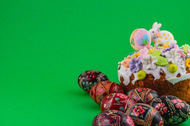 Süslü bir pasta parlak yeşil bir yüzeye oturur. Pastanın etrafında çeşitli renklerde boyanmış yumurtalar var. Sahne, özel bir gün için neşeli bir ruh hali sergiliyor..