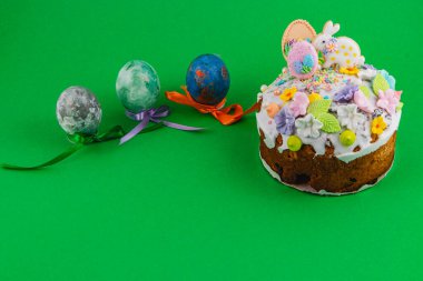 Kremalı ve süslemeli bir pasta, kurdeleli üç renkli yumurtanın yanında oturur. Bu sahne Paskalya şenlikleri sırasında parlak yeşil bir arkaplanda geçiyor..