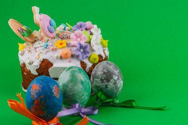 Paskalya pastası süslü yumurtaların yanına oturur. Pastanın kreması ve renkli süslemeleri var. Yumurtalar farklı renklerde ve kurdelelerle bağlanmış..