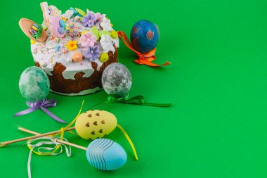 Krema ve çiçeklerle süslenmiş bir pasta parlak yeşil bir yüzey üzerinde renkli Paskalya yumurtalarının yanında oturur. Sahne, bayram için hazırlanan şenlikli eşyaları gösteriyor..