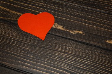 Kırmızı bir kalp şekli koyu ahşap bir yüzeye dayanır. Tahtanın kabataslak bir dokusu ve görünür tanecikleri var. Bu düzen aşkı veya arkadaşlığı sembolize edebilir..