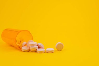 Bir şişe yan yatar, etrafına beyaz haplar saçılmış parlak sarı bir yüzeye. Bu sahne ilaçları basit bir ortamda gösteriyor..