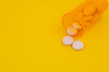 Haplar, bir konteynır düştükten sonra parlak sarı bir yüzeye saçılmış. Ayar turuncu bir şişenin yanında yuvarlak beyaz tabletlerin düzenlendiğini gösteriyor..