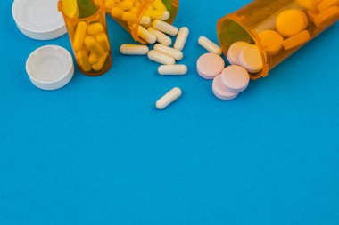 İçinde haplar olan şişeler mavi bir yüzeye dökülür. Farklı ilaç şekilleri ve renkleri etrafa saçılmış..