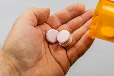 Bir el pembe hapları tutarken turuncu bir hap şişesi devriliyor. Sahne, ilaç alırken gelişigüzel bir an gösteriyor. Ayarlar sade ve eyleme odaklı..