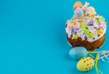 Paskalya için tasarlanmış bir pasta mavi bir yüzeyde bulunur. Üzerinde renkli süslemeler ve yakınlarda pastel renkli yumurtalar var. Sahne parlak ve şenlikli..