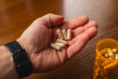 Bir insan elinde birden fazla beslenme kapsülü tutar. Ahşap bir masada daha fazla kapsül içeren bir şişenin yanında duruyorlar..