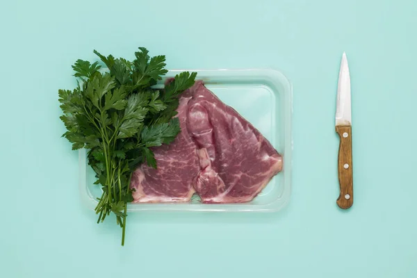 Uma faca com uma alça de madeira, um monte de salsa e carne em embalagem a vácuo em um fundo azul. — Fotografia de Stock