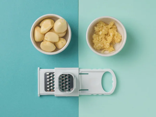 A bowl with whole and crushed garlic and a grater on a two-color background. A popular spice for the kitchen.