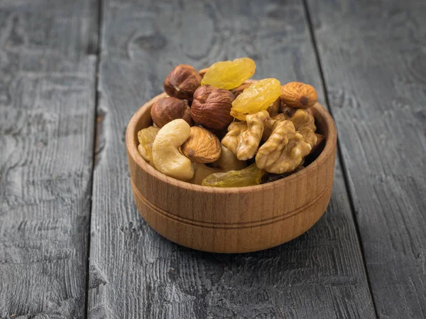 A wooden bowl with a mixture of nuts and dried fruits on a black wooden table. Natural healthy vegetarian food.