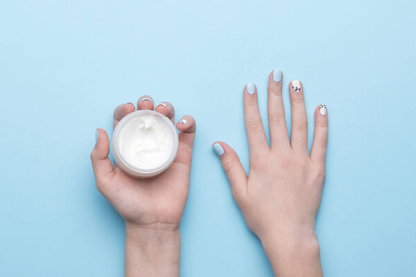 A jar of cream in women's hands with a stylish manicure on a blue background. Hand skin care. Flat lay.