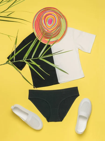 Black and white summer clothing set, colorful hat under the leaves of a palm tree on a yellow background. The concept of summer holidays.