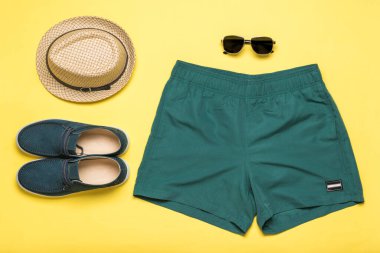 Sarı arka planda erkek ayakkabısı, şort, şapka ve gözlük. Popüler yaz erkek aksesuarları.