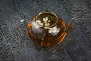 Tahta bir masanın üzerinde yasemin yaprakları ve çiçek çayı olan açık camdan bir çaydanlık. Sağlığın için iyi olan canlandırıcı bir içecek..