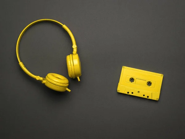 Classic yellow headphones and a yellow cassette with a magnetic tape on a black background. Color trend. Vintage equipment for listening to music. Flat lay.