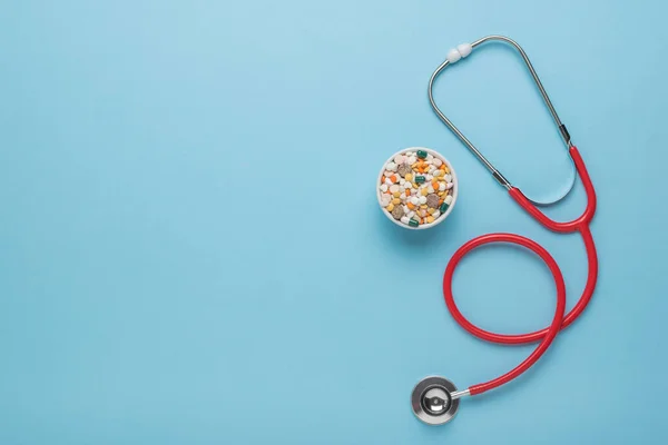 Medicines in a ceramic bowl and a red stethoscope on a blue background. Space for the text.The concept of treatment of various diseases. Flat lay.