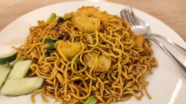 Yanında çatal ve kaşıkla servis edilen beyaz bir tabakta karides, sebze ve dilimlenmiş salatalıklı ramen tabağı. Asya mutfağı, lezzetli ve lezzetli yemek..