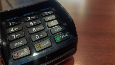 Tahta bir masanın üzerindeki kredi kartı ödeme terminalinin yakın çekimi elektronik işlemlerin tuş takımını gösteriyor. Masanın üzerinde nakitsiz ödemeler ve perakende işlemler için kullanılan bir satış noktası kart okuyucusu var.