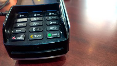 Tahta bir masanın üzerindeki kredi kartı ödeme terminalinin yakın çekimi elektronik işlemlerin tuş takımını gösteriyor. Masanın üzerinde nakitsiz ödemeler ve perakende işlemler için kullanılan bir satış noktası kart okuyucusu var.