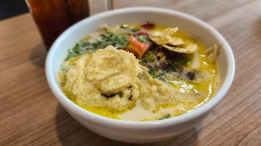 Bir kase geleneksel Endonezya soto çorbası. Zengin et suyu, hamur köftesi, taze otlar ve ahşap bir masada servis edilen baharatlar. Endonezya soto 'suna yakın plan, aromatik çorba, mantı, sebze ve taze garnitür..