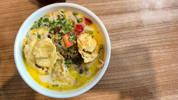 Bir kase geleneksel Endonezya soto çorbası. Zengin et suyu, hamur köftesi, taze otlar ve ahşap bir masada servis edilen baharatlar. Endonezya soto 'suna yakın plan, aromatik çorba, mantı, sebze ve taze garnitür..