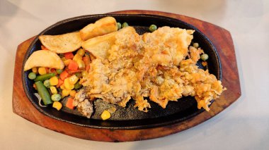 Kızarmış tavuk bifteği, karışık sebzeler ve patates dilimleriyle servis edilir. Lezzetli kızarmış tavuk filetosu ve renkli sebzeler, cızırdayan siyah bir tabakta..