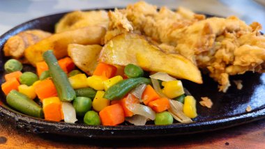 Kızarmış tavuk bifteği, karışık sebzeler ve patates dilimleriyle servis edilir. Lezzetli kızarmış tavuk filetosu ve renkli sebzeler, cızırdayan siyah bir tabakta..