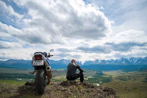 Glatzköpfiger sitzt auf einem Berg neben einem Motorrad und sieht aus. Stockbild