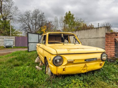 Vinnytsia, Ukrayna - 23 Ekim 2025: Sonbahar ağaçları ve bulutlu gökyüzü ile bir banliyö bahçesine atılmış eski sarı ZAZ-968 model bir arabanın ön görüntüsü. Yüksek kalite fotoğraf